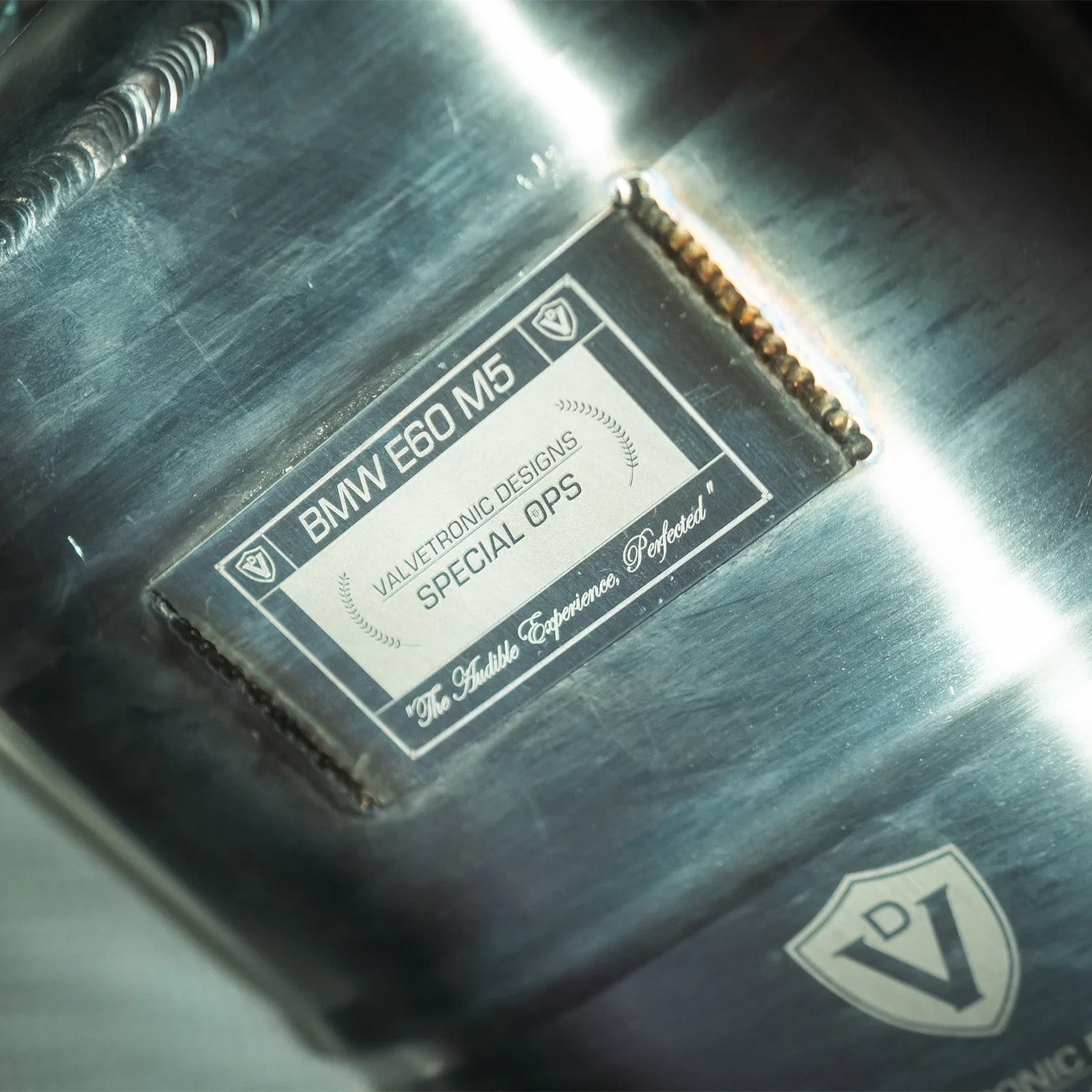 Close-up of BMW E60 M5 Valvetronic exhaust with a label and logo, featuring 'Valvetronic Designs' and 'BMW E60 M5'.
